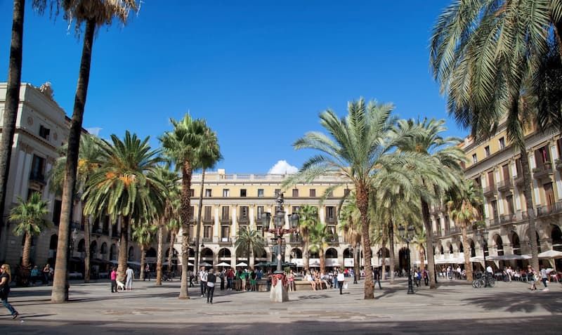 Fotografia di Plaça Reial di Barcellona con cielo sereno, palme piantate a terra nel cemento e persone che camminano