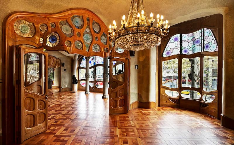 Fotografia dell'interno di Casa Milà di Barcellona con parquet in legno, candeliere con luci accese e decorazioni in legno alle porte e finestre