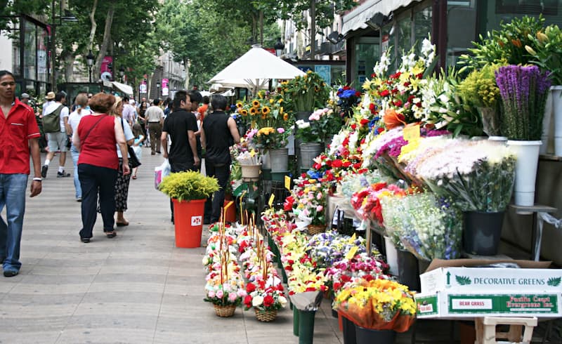 Fotografia di una bancarella che vende fiori sulla destra con persone che camminano sul lato sinistro della Rambla de Les Flors di Barcellona