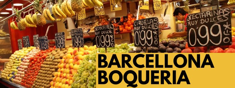 Fotografia di una bancaerella di frutta con i prezzi esposti e rettangolo arancione in basso a destra con scritta nera Barcellona Boqueria