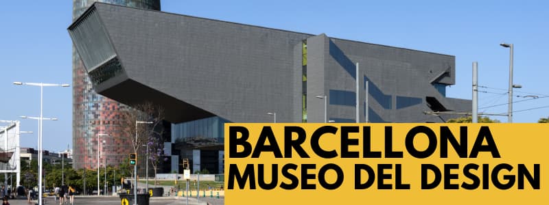 Fotografia dall'esterno del Museo del Design di Barcellona con struttura a forma di parallelepipedo con una torre accanto con cielo azzurro alle spalle. In basso a destra rettangolo arancione con scritta nera "Barcellona museo del design"