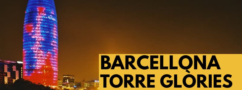 Fotografia della Torre Glòries di Barcellona di notte, un grattacielo illuminato blu, viola e rosso sulla sinistra con gli altri edifici in basso. In basso a destra rettangolo arancione con scritta nera "Barcellona Torre Glòries"